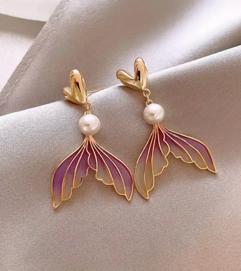 Pendientes dorados con perla y colgante de ala de mariposa en tonos rosa y lila sobre tela gris