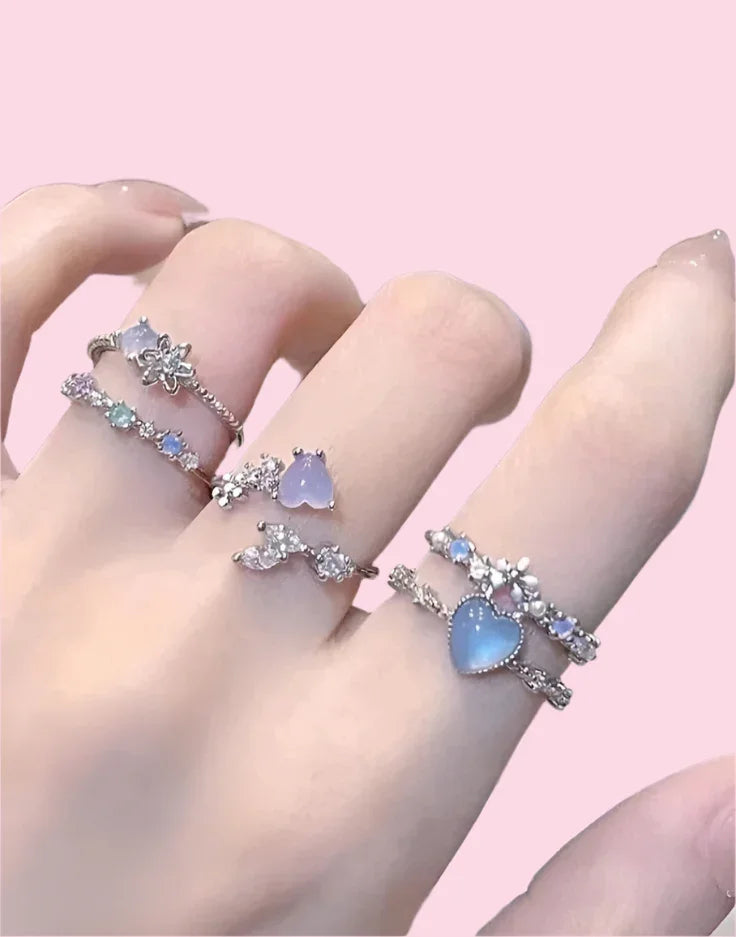 Mano con anillos plateados coreanos con piedras azules y decoraciones florales sobre fondo rosa.