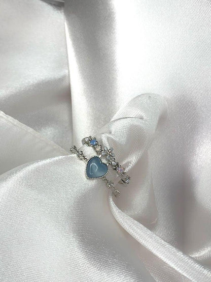 Anillo de plata con corazón azul y detalles de flores sobre tela satinada blanca.