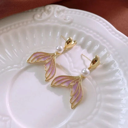 Aretes dorados en forma de cola de sirena con detalles rosados y perla, sobre plato blanco